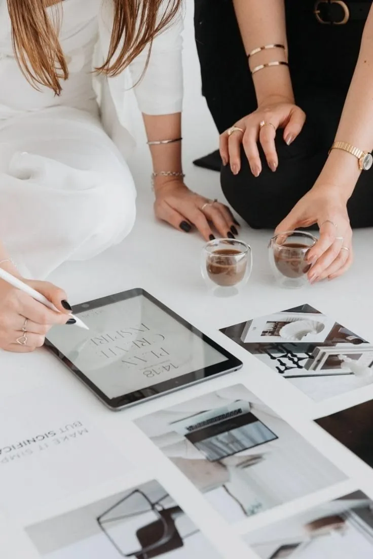 Two women planning content on a tablet over a moodboard of printed photos and coffee